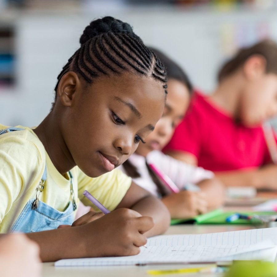 Cute pupil writing at desk in classroom at the elementary school. Student girl doing test in primary school. Children writing notes in classroom. African schoolgirl writing on notebook during the lesson.